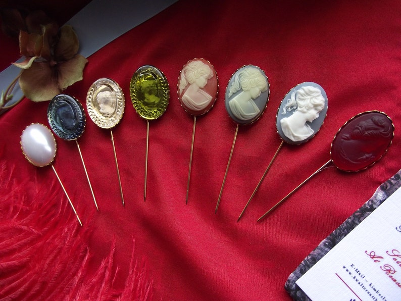 May include: Assortment of eight vintage cameo brooches with gold-tone settings. The cameos feature various designs, including a woman's profile, a floral pattern, and a geometric design. The brooches are displayed on a red fabric background.