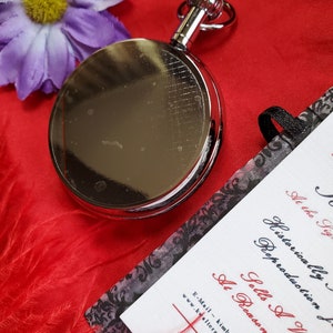 May include: A shiny silver pocket watch with a ring at the top, resting on a vibrant red surface. A purple flower and a card with black and red text are also in the image. The card reads "Historically Reproduction Sells A Variety At Reasonable Ra..."