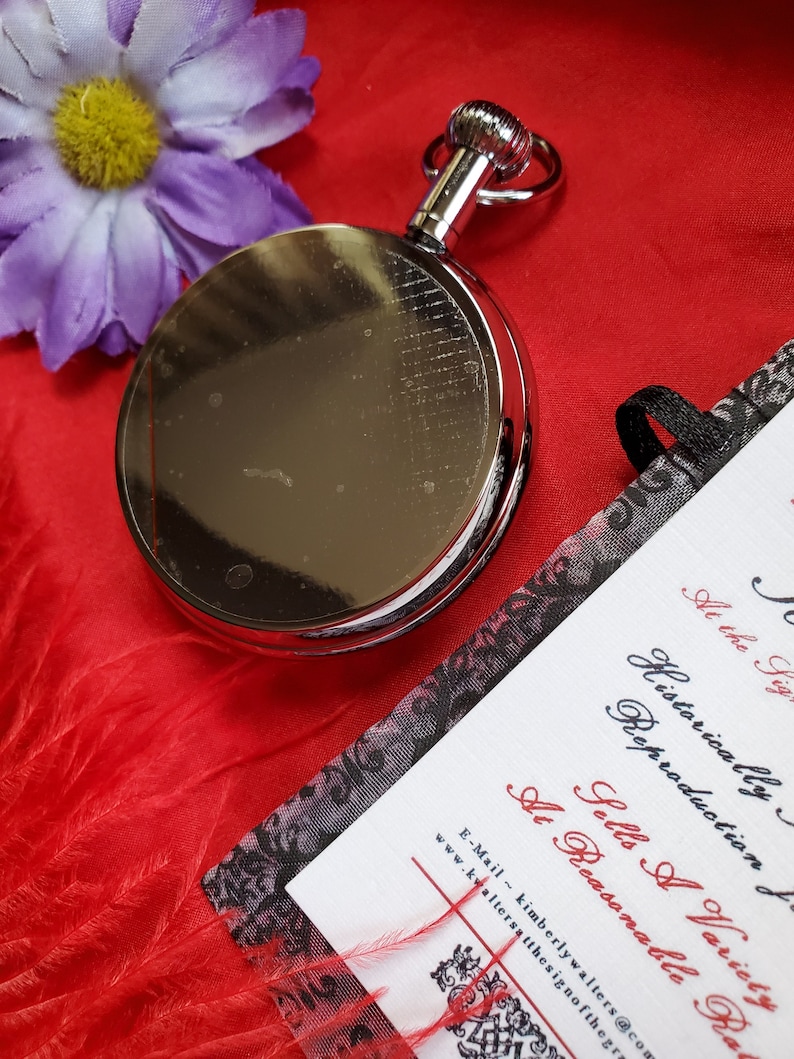May include: A shiny silver pocket watch with a ring at the top, resting on a vibrant red surface. A purple flower and a card with black and red text are also in the image. The card reads "Historically Reproduction Sells A Variety At Reasonable Ra..."