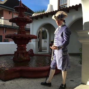 May include: A person in a historical costume stands near a red fountain. The costume includes a light purple jacket and breeches, a white shirt, and a tricorn hat. The person is holding a cane and wearing black shoes.