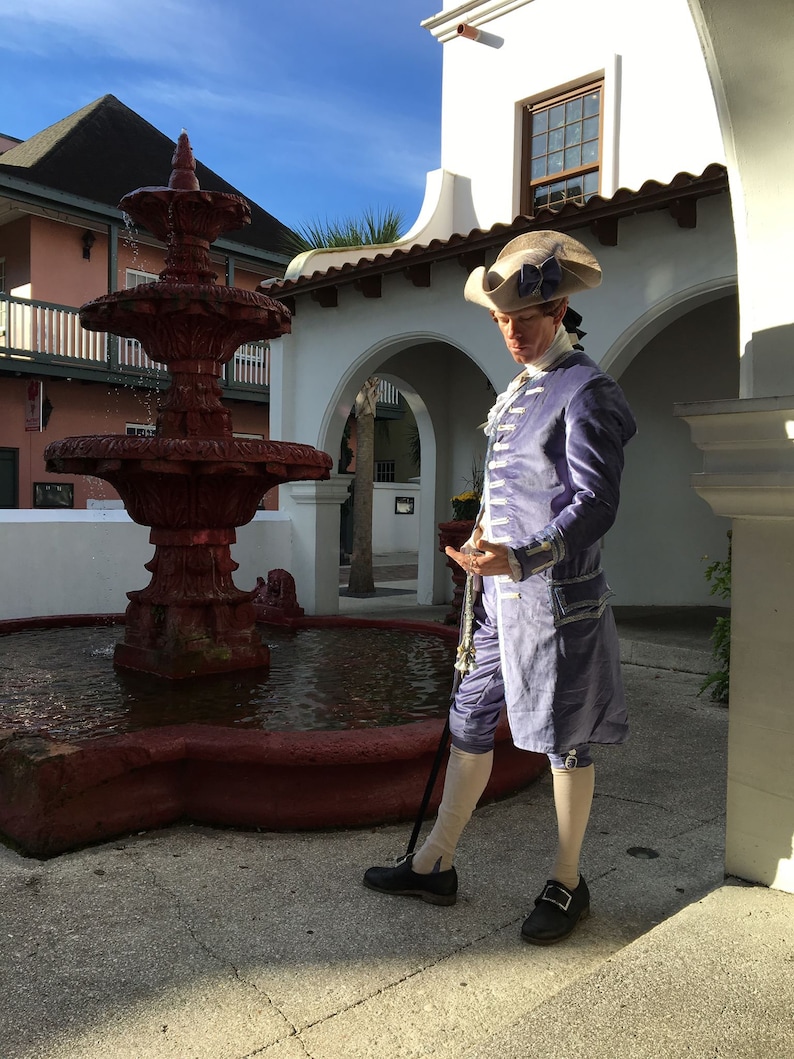May include: A person in a historical costume stands near a red fountain. The costume includes a light purple jacket and breeches, a white shirt, and a tricorn hat. The person is holding a cane and wearing black shoes.