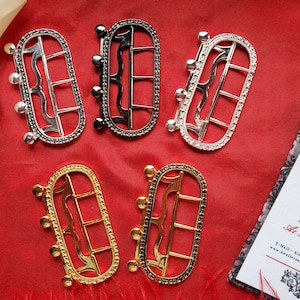 Puede incluir: Cinco hebillas de cinturón de metal adornadas con diferentes acabados. Las hebillas son de forma ovalada con un diseño decorativo y tienen tres pequeños remaches redondos a cada lado. Las hebillas son plateadas, negras, doradas y dos tonos diferentes de plata.