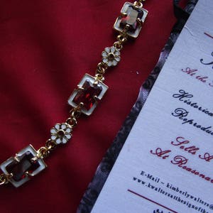 May include: A gold bracelet with white flower accents and red gemstone accents. The bracelet is on a red fabric background.