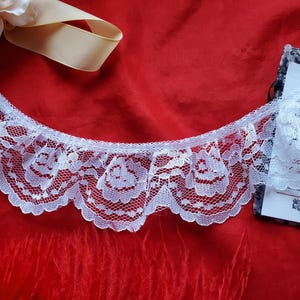 Puede incluir: Ribete de encaje blanco con borde festoneado y diseño floral, sobre tela roja. También hay una cinta color crema y una pequeña tarjeta con texto. El encaje es delicado y detallado.