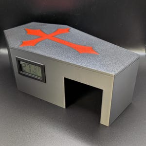 May include: A gray coffin-shaped box with a red cross on the lid. The box has a digital display showing numbers and a rectangular opening on the side. The surface has a textured appearance.