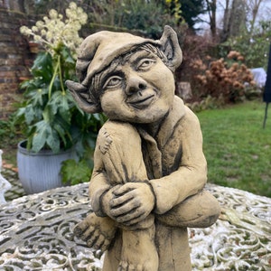 Puede incluir: Una escultura de gnomo de jardín caprichosa hecha de piedra, con un sombrero puntiagudo, orejas grandes y una pose sentada. El gnomo está pintado de color marrón claro y tiene una expresión amigable.
