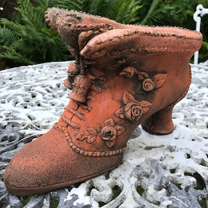 Peut inclure: Jardinière en terre cuite en forme de chaussure brune avec des détails floraux. La chaussure a un talon haut et un laçage décoratif. La jardinière est décorée de roses et de feuilles.