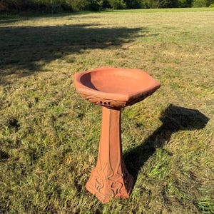 May include: A terracotta birdbath featuring a wide, shallow basin with a hexagonal rim and a tall, tapered pedestal base. The exterior of the pedestal and the underside of the basin are decorated with raised, flowing, organic, leaf-like patterns.