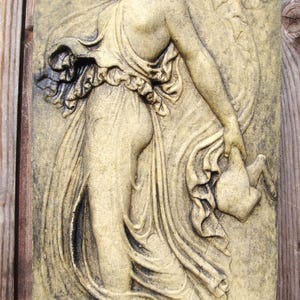 May include: A rectangular, weathered, beige relief sculpture of a classical figure in flowing robes, holding a jug. The figure is depicted in a walking pose, with detailed drapery and a serene expression. The sculpture is mounted on a wooden background.