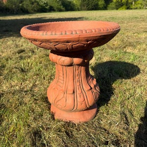 May include: A terracotta birdbath featuring a wide, circular basin with a decorative scalloped rim. The pedestal base is cylindrical with vertical fluted carvings and rounded accents, showcasing a natural reddish-brown clay finish throughout.
