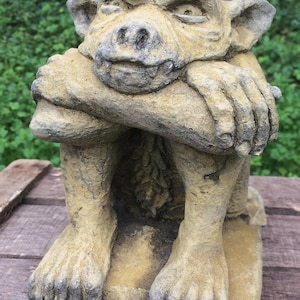 May include: A weathered, beige stone gargoyle statue featuring a squatting creature with pointed ears, a flat nose, and a wide mouth. The figure is depicted with its arms crossed, resting its chin on its forearms, showcasing a textured surface.