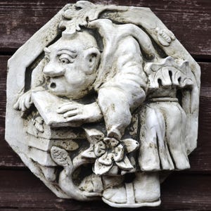 Op de afbeelding: Een witte, verweerde stenen wandplaat met een reliëfsculptuur van een figuur met een groot hoofd en wijde ogen die over een rechthoekige structuur leunt. De figuur is omringd door uitgesneden florale elementen, bladeren en een klein insect.