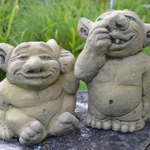 Op de afbeelding: Twee stenen tuinkabouters, een bedekt zijn ogen en de ander bedekt zijn oren. Beide kabouters lachen en hebben een grillige, speelse uitdrukking.