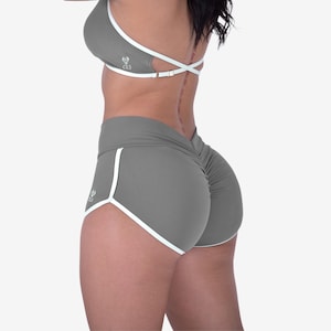May include: Grey athletic shorts and sports bra set with white trim. The shorts have a ruched back and a small logo on the side. The sports bra has a criss-cross back design. The set is made of a stretchy material.