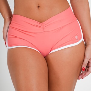May include: Coral-colored athletic shorts featuring a V-shaped waistband with subtle center ruching and white trim along the curved leg openings. A small white logo is visible on the lower left side of the shorts, which are designed with a fitted silhouette.