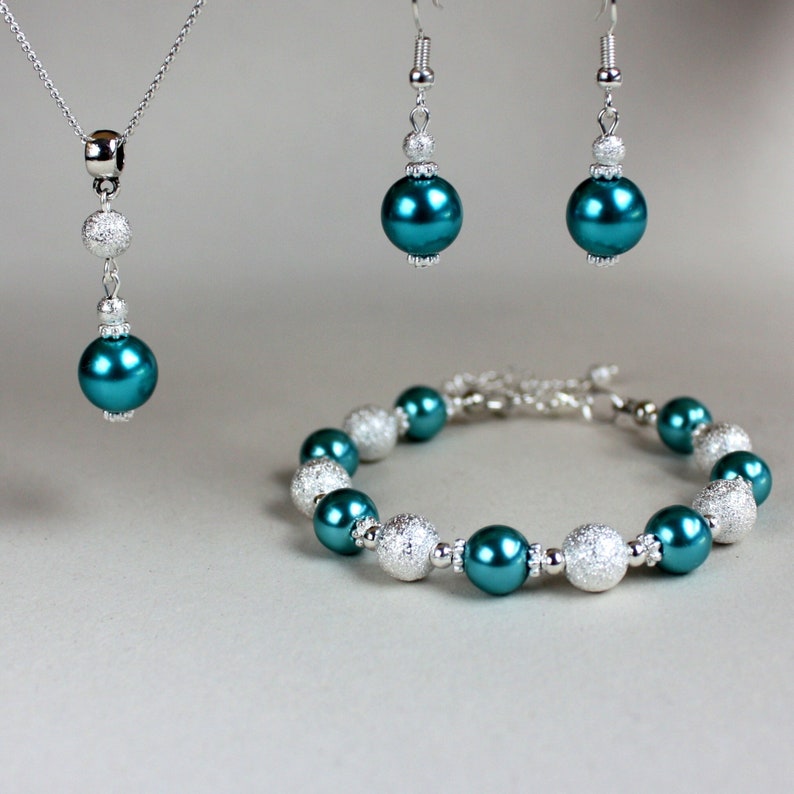 May include: Teal and silver bead jewelry set including a necklace, earrings, and bracelet. The necklace has a single large teal bead and two smaller silver beads. The earrings have a single large teal bead and a smaller silver bead. The bracelet has alternating teal and silver beads.