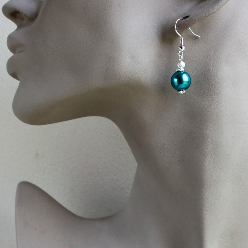 May include: Teal pearl dangle earrings with silver accents. The earrings feature a single large teal pearl bead and smaller white beads on the silver chain.