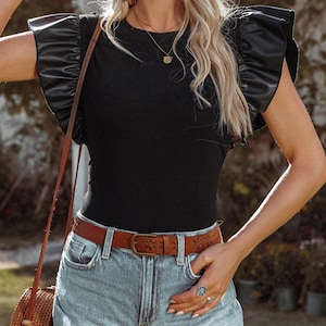 May include: A black sleeveless top with a round neckline and faux leather ruffle sleeves. The top is paired with a brown belt with a floral design and light blue denim shorts.