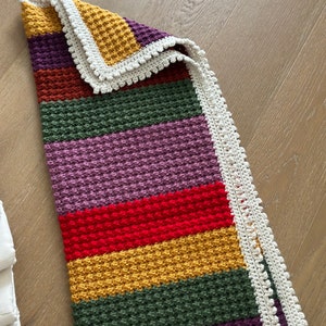 May include: A multi-colored crocheted baby blanket with a white crocheted border. The blanket is made with a textured stitch and features stripes of purple, green, red, yellow, and brown.
