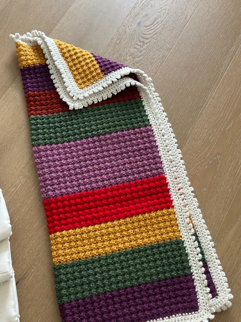 May include: A multi-colored crocheted baby blanket with a white crocheted border. The blanket is made with a textured stitch and features stripes of purple, green, red, yellow, and brown.