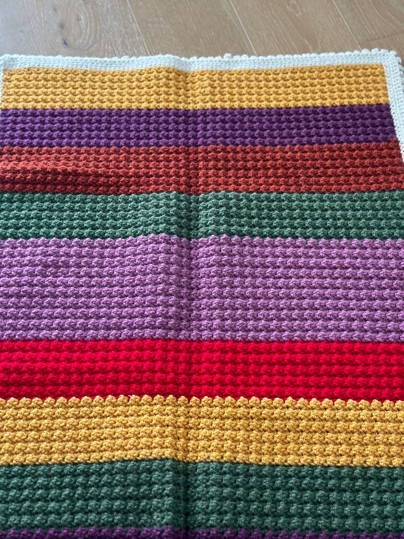 May include: A multi-colored crocheted blanket with stripes of yellow, purple, brown, green, lavender, red, and green. The blanket is made with a textured stitch.