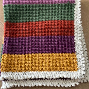 May include: A crocheted baby blanket with a textured waffle stitch in shades of purple, green, orange, and yellow. The blanket has a white crocheted border.