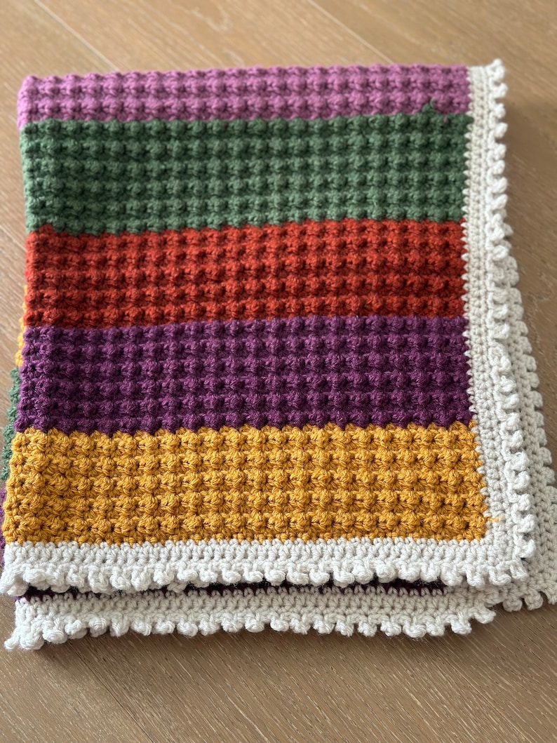May include: A crocheted baby blanket with a textured waffle stitch in shades of purple, green, orange, and yellow. The blanket has a white crocheted border.