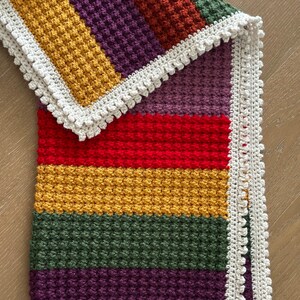 May include: A multi-colored crocheted baby blanket with a white crocheted border. The blanket is made with a textured stitch and features stripes of red, yellow, green, purple, and orange.