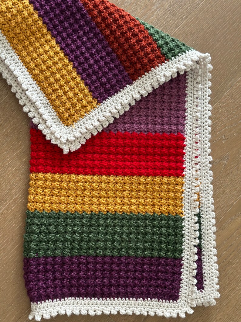 May include: A multi-colored crocheted baby blanket with a white crocheted border. The blanket is made with a textured stitch and features stripes of red, yellow, green, purple, and orange.