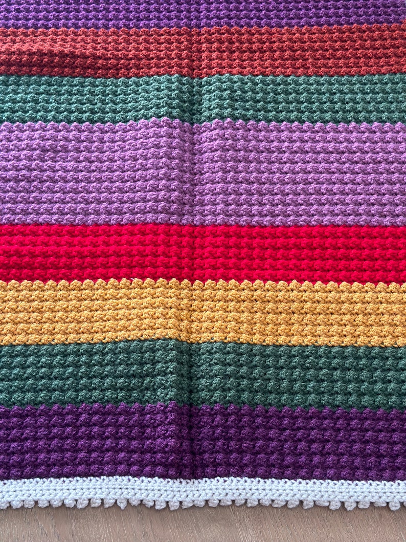 May include: A multi-colored crocheted blanket with a white crocheted border. The blanket is made with a textured stitch and features stripes of purple, brown, green, red, yellow, and dark purple.