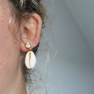 Op de afbeelding: Een paar gouden oorbellen met een witte schelpbedel en een heldere strasssteen. De oorbellen worden gedragen door een persoon met blond haar.