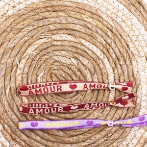 Op de afbeelding: Drie kleurrijke vriendschapsarmbanden met het woord "Amour" en een hartvormig ontwerp. De armbanden liggen op een geweven stroomachtige achtergrond.