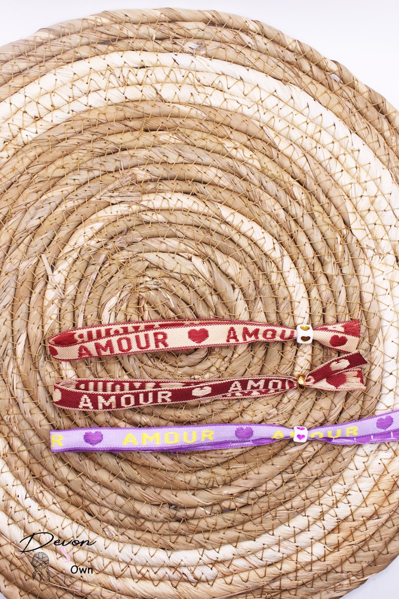 Op de afbeelding: Drie kleurrijke vriendschapsarmbanden met het woord "Amour" en een hartvormig ontwerp. De armbanden liggen op een geweven stroomachtige achtergrond.