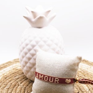 Op de afbeelding: Een wit keramiek ananasbeeldje staat op een geweven stro mat. Ernaast ligt een klein, cr&egrave;mekleurig kussen met een rood en gouden armbandje waarop "AMOUR" staat met een klein hartje.