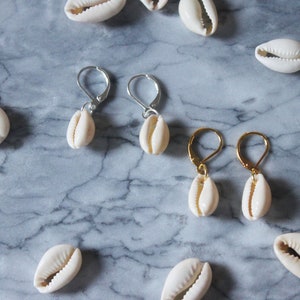 May include: Two pairs of earrings with cowrie shells hanging from silver and gold hooks. The shells are white and the hooks are silver and gold. The earrings are on a marble surface.
