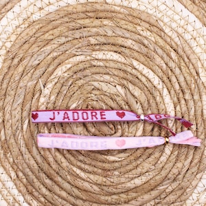 Op de afbeelding: Twee roze en rode vriendschapsarmbanden met de tekst "J'ADORE" en een klein hartje op elke armband. De armbanden liggen op een geweven strooien achtergrond.