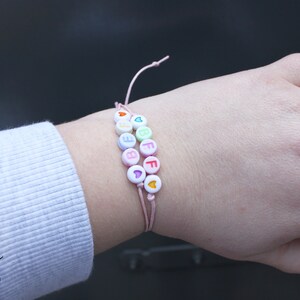 May include: A pink string bracelet with white beads that spell out "BFF" with colorful hearts in between the letters.