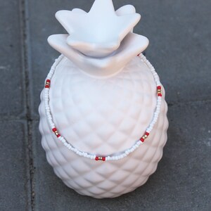 May include: White and red beaded necklace with gold accents. The necklace is displayed on a white ceramic pineapple.