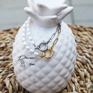 May include: A white pearl necklace and a silver chain with a gold clasp are displayed on a white pineapple-shaped ceramic object. The necklace and chain are layered on top of each other. The text "Devon & Own" is visible on the ceramic object.