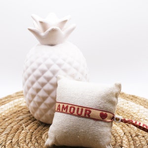 Op de afbeelding: Een klein wit kussen met een rood en beige lint waarop "AMOUR" staat met een rood hartje erop. Het kussen ligt op een geweven stro mat met een witte keramische ananas erachter.