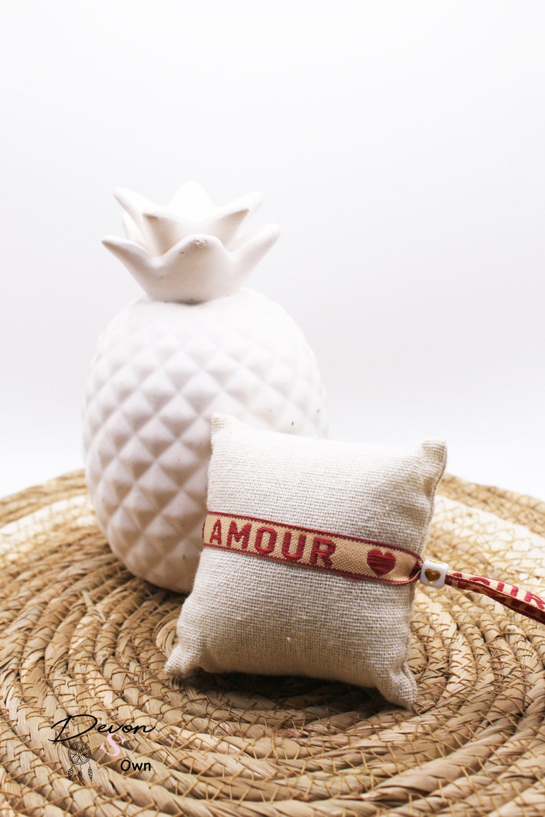 Op de afbeelding: Een klein wit kussen met een rood en beige lint waarop "AMOUR" staat met een rood hartje erop. Het kussen ligt op een geweven stro mat met een witte keramische ananas erachter.
