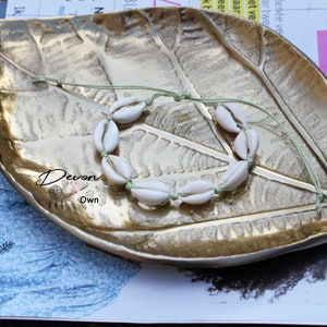 May include: A white shell bracelet with a green string on a gold leaf-shaped tray. The bracelet is made of small, white seashells strung together with a green cord.