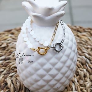 May include: A white heart-shaped bead bracelet with a gold and silver clasp. The bracelet is displayed on a white pineapple-shaped stand.