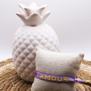 Op de afbeelding: Een klein, wit keramiek ananasbeeldje staat op een geweven strooien mat. Ernaast ligt een klein, beige stoffen zakje met een paars lint waarop in gele letters "AMOUR" staat.