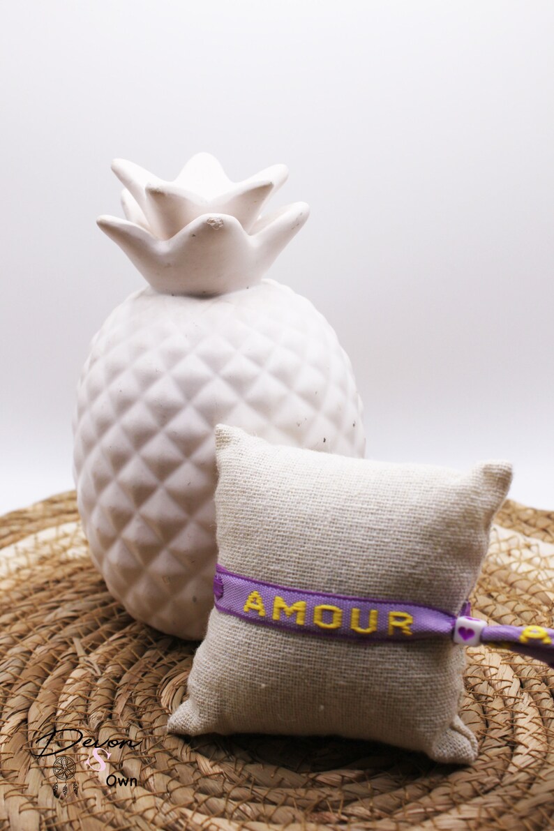 Op de afbeelding: Een klein, wit keramiek ananasbeeldje staat op een geweven strooien mat. Ernaast ligt een klein, beige stoffen zakje met een paars lint waarop in gele letters "AMOUR" staat.