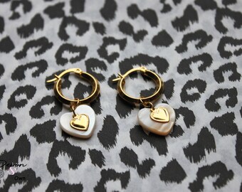Puede incluir: Aretes de aro dorados con un dije de corazón blanco que presenta un corazón dorado más pequeño en el centro. Los aretes están sobre un fondo de estampado de leopardo blanco y negro.