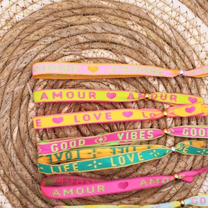 Op de afbeelding: Een set kleurrijke geweven vriendschapsarmbanden met verschillende zinnen en harten. De armbanden bevatten zinnen zoals "Amour", "Love", "Good Vibes" en "Life Love".
