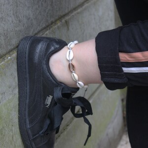 Op de afbeelding: Een witte en gouden schelpen enkelband met een delicate ketting. De enkelband wordt gedragen op de enkel van een persoon, met een zwarte sneaker en een zwarte broek met witte en bruine strepen zichtbaar.