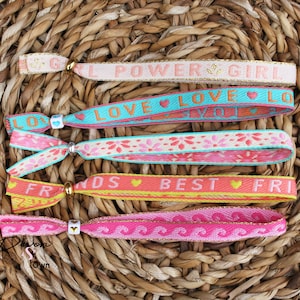 Op de afbeelding: Vijf kleurrijke vriendschapsarmbanden met verschillende boodschappen, waaronder "Girl Power", "Love", "Friends" en "Best Friends". De armbanden zijn gemaakt van geweven stof en hebben kleine hartcharms.