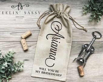 May include: A burlap wine bottle bag with the text "Will you be my bridesmaid?" and the name "Ariana" written in cursive. The bag is on a wooden surface with a corkscrew and wine corks.